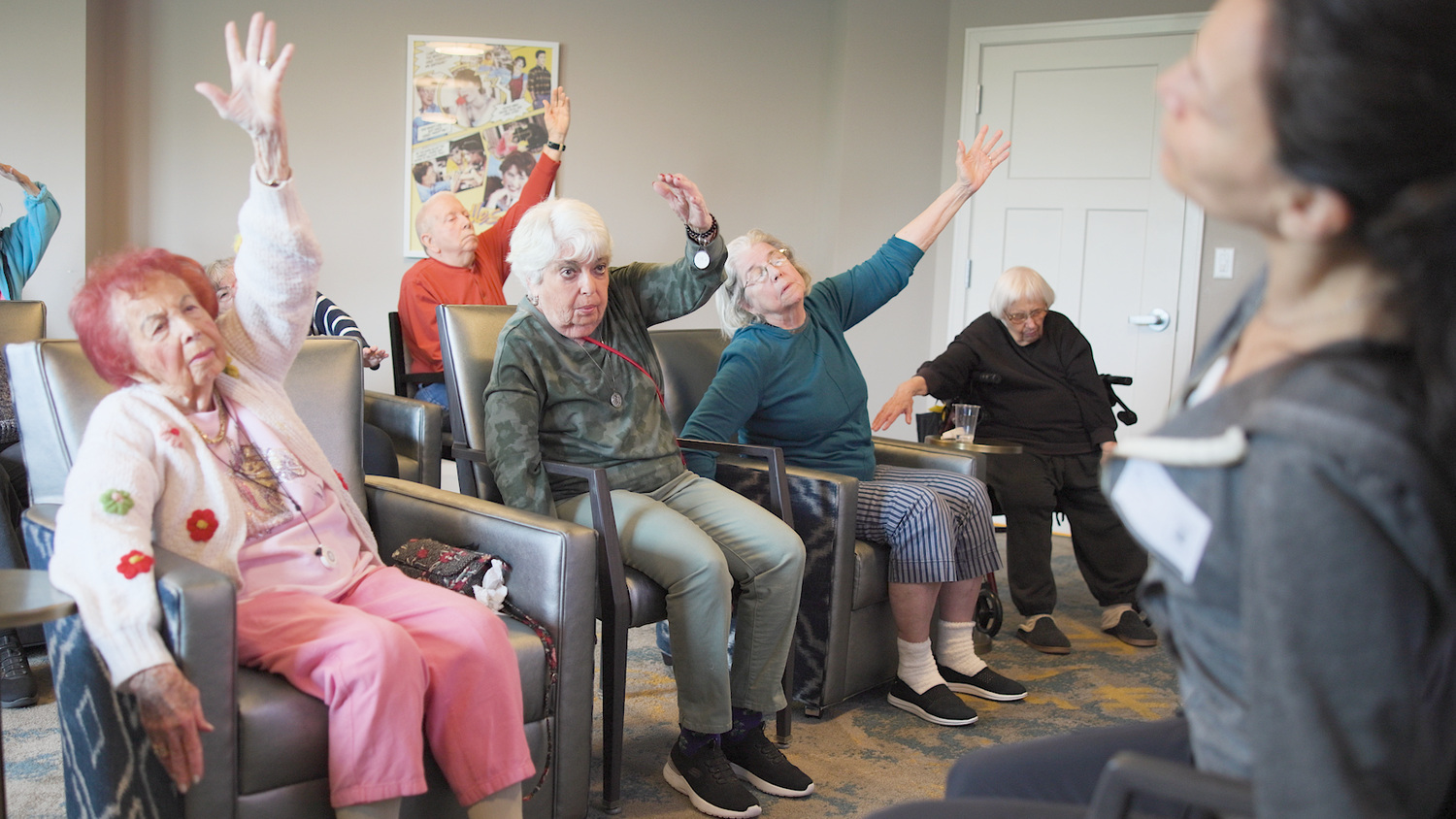 A group of residents raises their arms together during a seated stretching class in the home cinema room, guided by a staff member nearby, part of the active senior lifestyle Highland Park IL residents enjoy at our senior living Highland Park IL community.