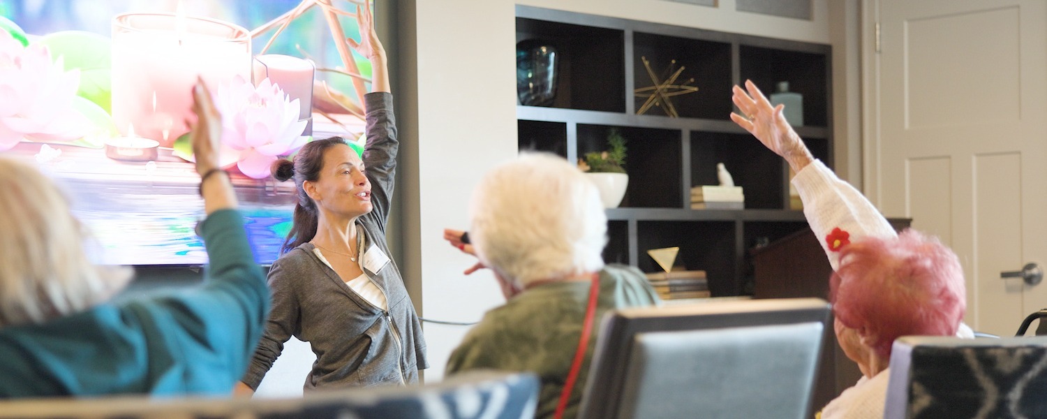 A woman at the front of the room raises her arm overhead as seated community members follow along, stretching near a screen showing pink lotus flowers at Arbor Terrace Highland Park, a calming moment of wellness-focused senior living North Shore and an active senior lifestyle Highland Park IL.