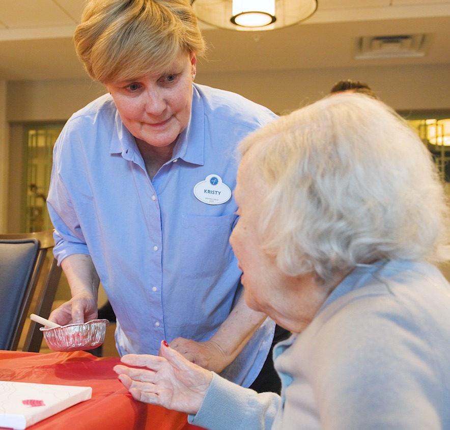 A woman in a light blue shirt holds a small foil dish with a brush as she leans in to chat with a community member beside a canvas on a red table at Arbor Terrace Highland Park, a warm, creative moment of personalized senior care Highland Park IL and an active senior lifestyle Highland Park IL.