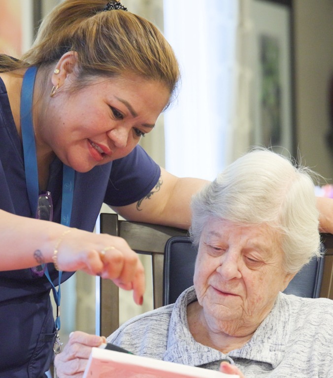 A woman in a navy top leans in to help a community member with a colorful project at a table at Arbor Terrace Highland Park, a warm, attentive moment that reflects personalized senior care Highland Park IL and friendly senior living Highland Park IL.
