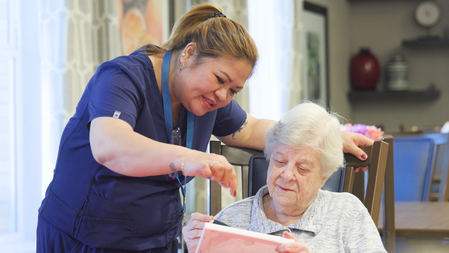 A smiling member of the senior living team helps a resident navigate a tablet together, capturing a warm, everyday moment of connection within this senior living community.