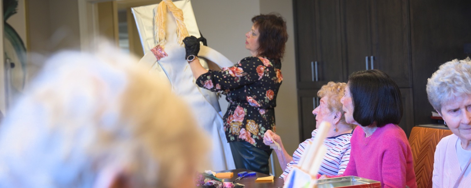 A woman in a floral blouse adds fabric details to a large figure with braided yarn hair as community members watch from a table of craft supplies at Arbor Terrace Highland Park, one of the top senior living communities Lake County IL, celebrating an active senior lifestyle Highland Park IL.