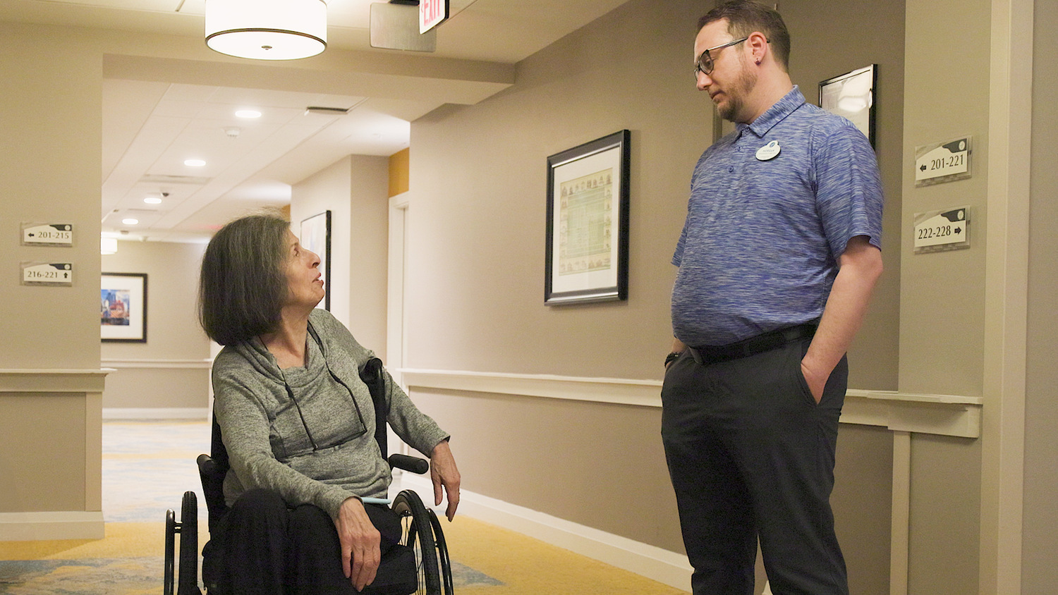 A staff member chats warmly with a resident in a bright hallway lined with framed artwork, sharing a friendly moment at our senior living Highland Park IL community and its active senior lifestyle Highland Park IL.