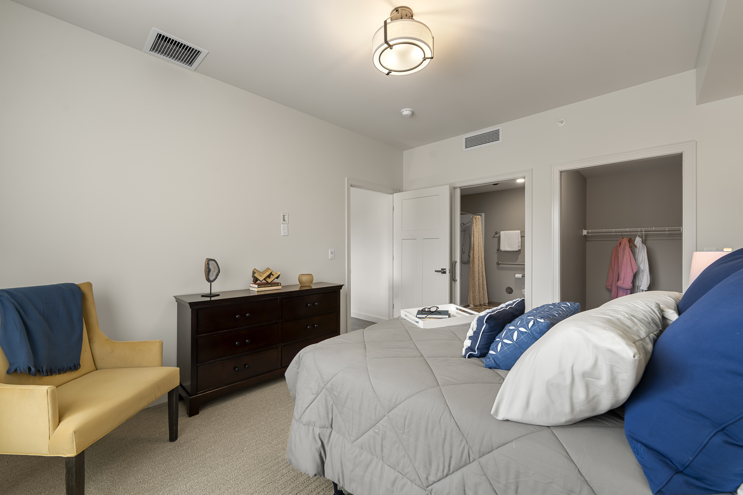 A spacious bedroom features a gray bedspread with navy accent pillows, a wood dresser, and a mustard yellow accent chair, with a closet and en-suite bathroom nearby, showcasing an apartment at our senior living Highland Park IL community and its active senior lifestyle Highland Park IL.