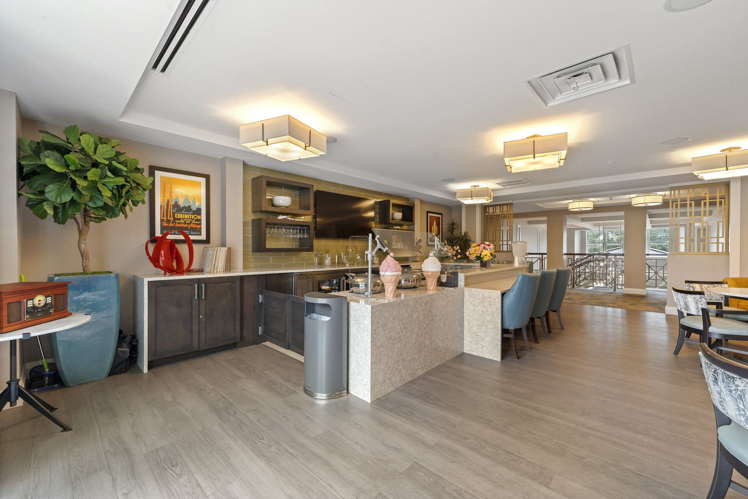 A cozy bistro area features a marble kitchen island with playful ice cream cone decor, blue upholstered chairs, a potted plant, and a vintage radio nearby, showcasing the wellness-focused senior living North Shore amenities at our senior living Highland Park IL community.
