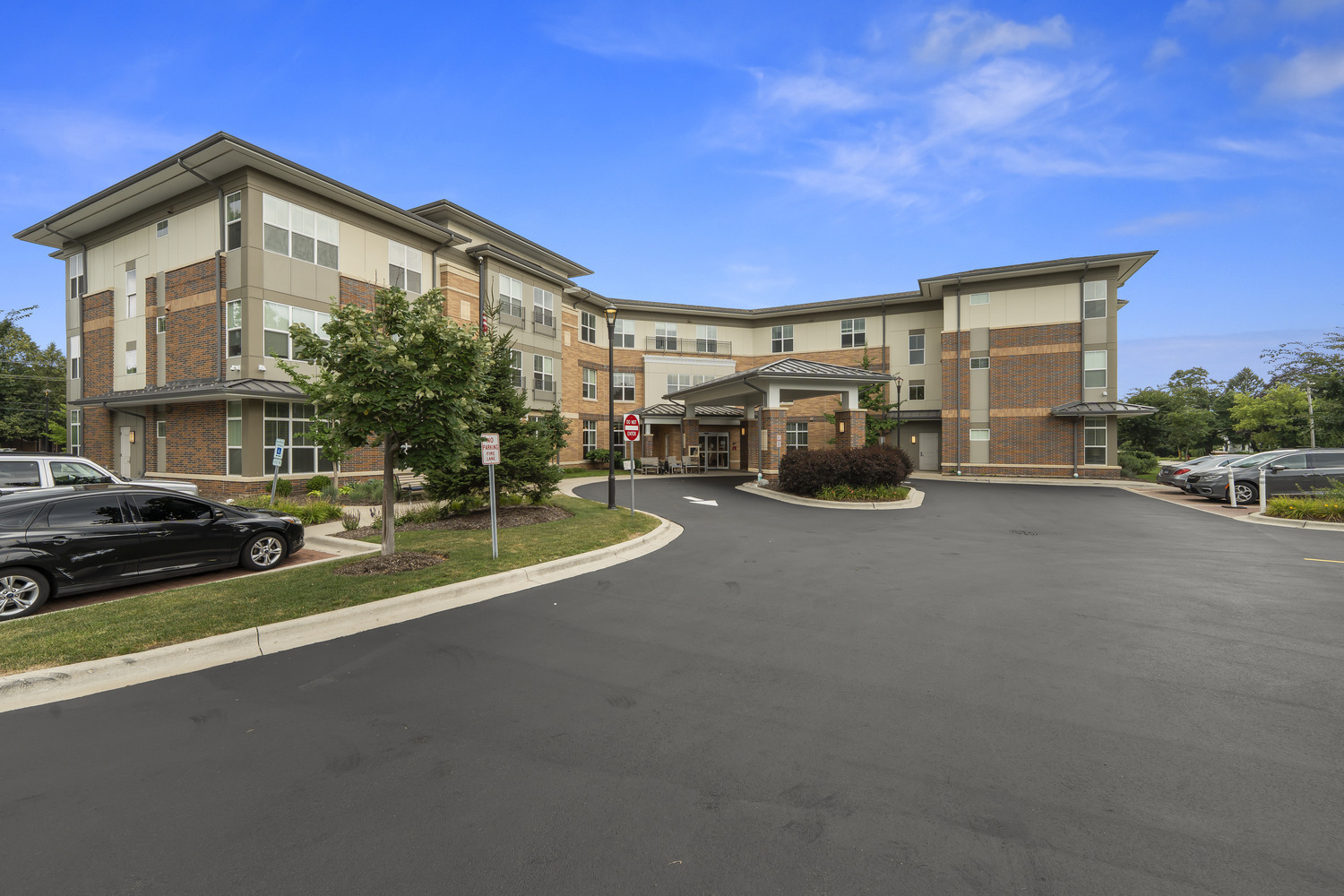A wide exterior view showcases the brick and stone building of our senior living Highland Park IL community, featuring a covered entrance, parking area, and landscaped grounds beneath a bright blue sky, reflecting the active senior lifestyle Highland Park IL residents enjoy.