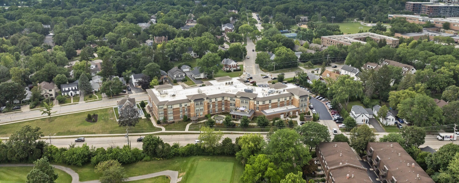 Bird's-eye view of a brick community surrounded by mature trees, neighborhood homes, and an open green lawn with a winding path, highlighting senior living with walking paths Highland Park IL and a welcoming setting for senior care Highland Park Illinois.