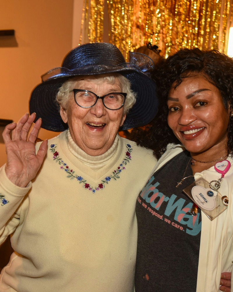 A laughing community member in a navy hat and cream sweater with embroidered flowers waves beside a smiling woman by a shimmering gold backdrop at Arbor Terrace Highland Park, one of the top senior living communities Lake County IL, offering warm senior care Highland Park Illinois.