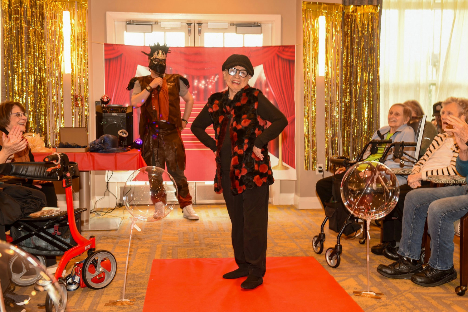A resident strikes a confident pose on a red carpet runway during a lively fashion show, as an entertainer hosts and fellow residents applaud from their seats, reflecting the active senior lifestyle Highland Park IL residents enjoy at our senior living Highland Park IL community.