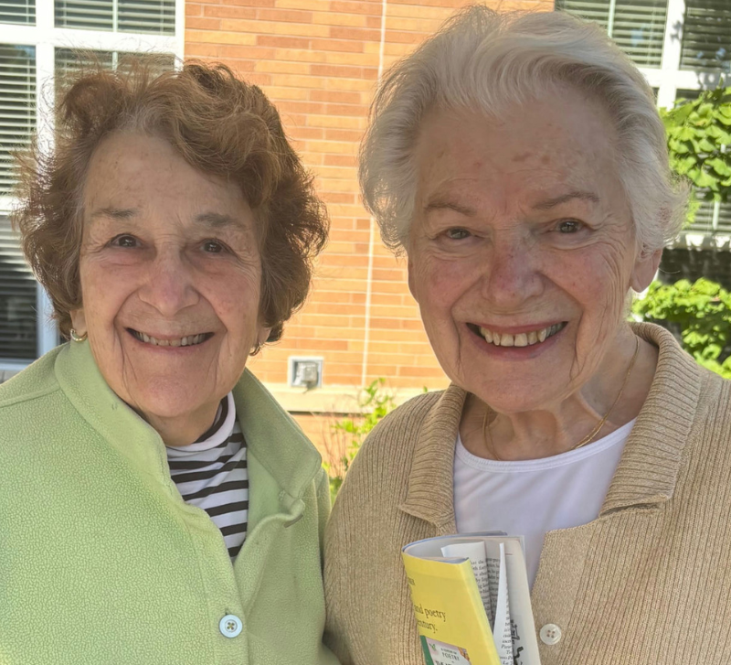 Two smiling community members stand close together in the sunshine, one in a soft green jacket and the other in a tan cardigan holding an open book, sharing a bright moment at Arbor Terrace Highland Park that captures the active senior lifestyle Highland Park IL and senior living Highland Park IL.