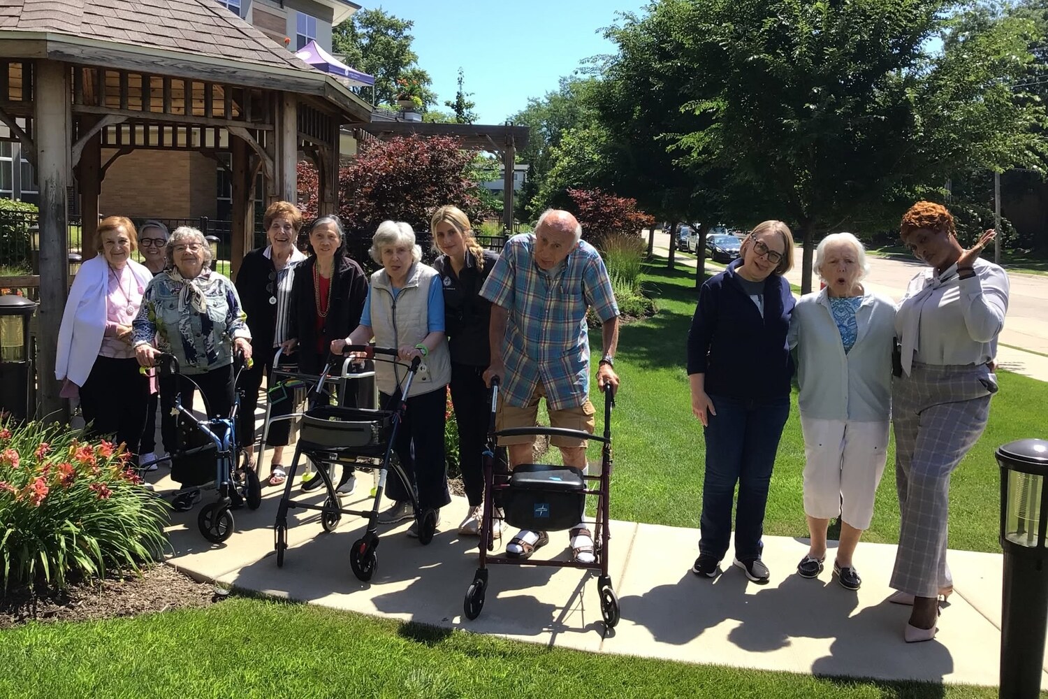 A group gathers on a sunny walkway beside a wooden gazebo, green lawn, and leafy trees at Arbor Terrace Highland Park, a bright outdoor moment of senior living with walking paths Highland Park IL and nature-friendly senior living Highland Park IL.