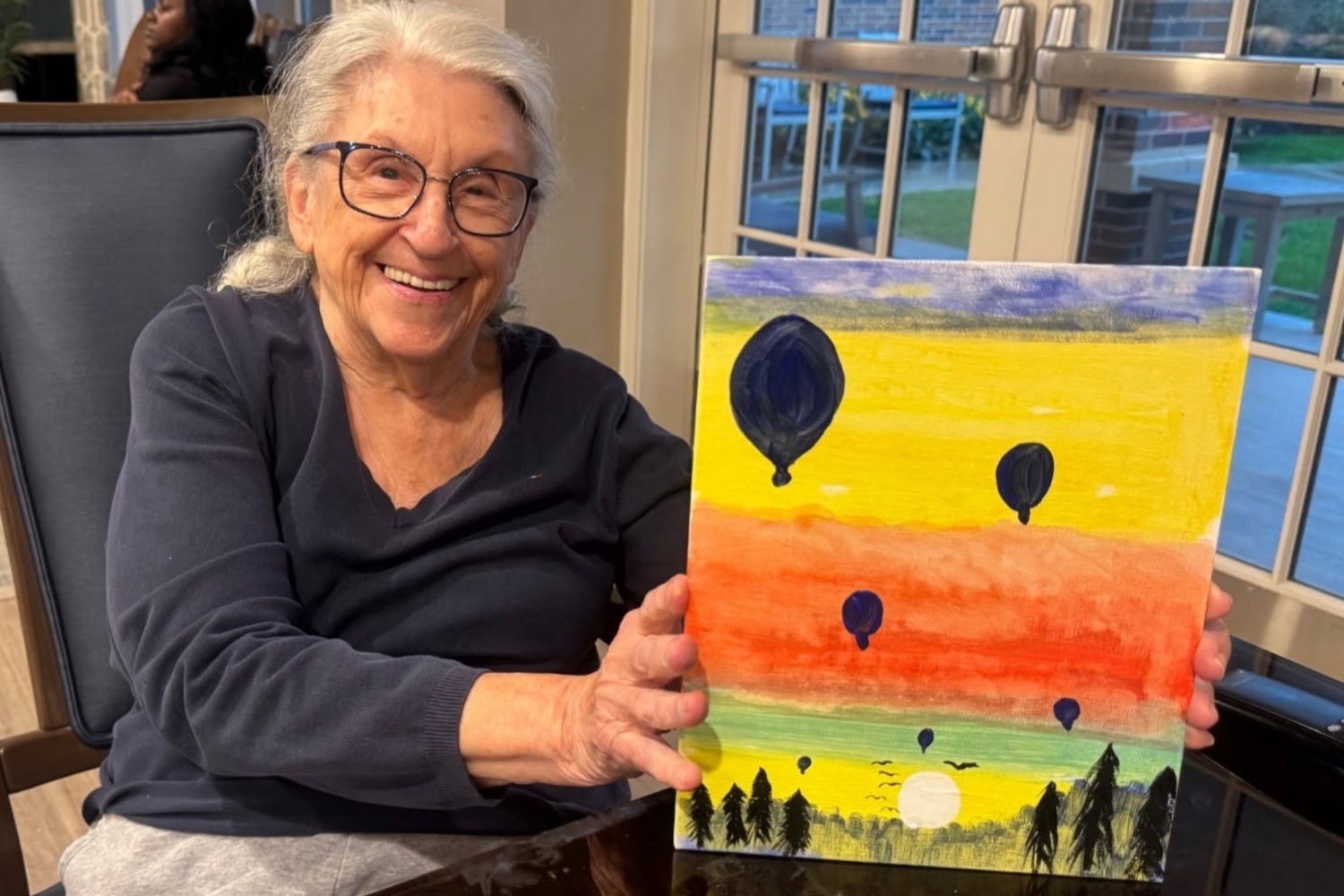 A smiling community member in glasses proudly holds up her painting of hot air balloons floating over a glowing yellow and orange sky above silhouetted trees at Arbor Terrace Highland Park, celebrating creativity in senior living Highland Park IL and an active senior lifestyle Highland Park IL.