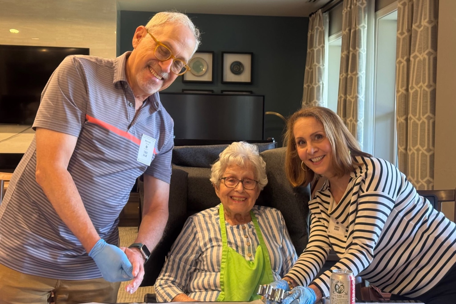 A smiling community member in a green apron sits at a table between a man in a striped polo and a woman in a black and white striped top, gathered for a hands-on activity at Arbor Terrace Highland Park that reflects senior living Highland Park IL and personalized senior care Highland Park IL.
