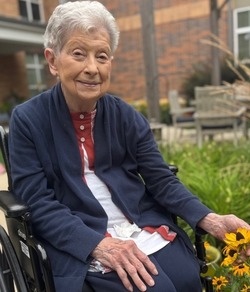 A smiling community member in a navy cardigan rests her hand near bright yellow flowers in a garden with a brick building and patio chairs behind her at Arbor Terrace Highland Park, a peaceful outdoor moment of nature-friendly senior living Highland Park IL and senior care Highland Park Illinois.
