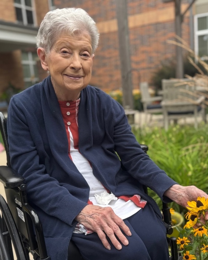A smiling community member in a navy cardigan rests her hand near bright yellow flowers in a garden with a brick building and patio chairs behind her at Arbor Terrace Highland Park, a peaceful outdoor moment of nature-friendly senior living Highland Park IL and senior care Highland Park Illinois.