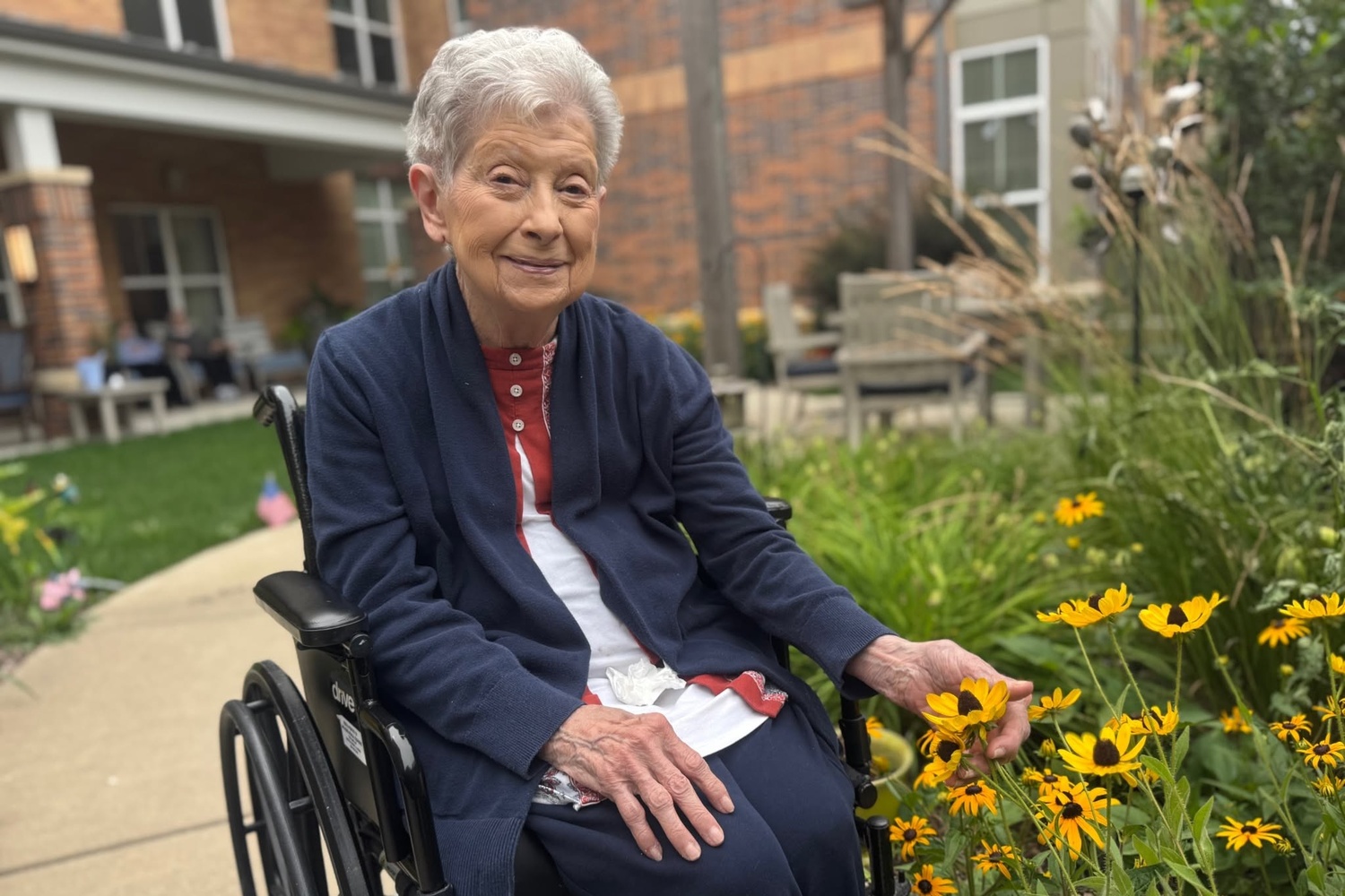 A smiling community member in a navy cardigan reaches toward bright yellow flowers in a garden bed beside a walkway at Arbor Terrace Highland Park, a peaceful outdoor moment of nature-friendly senior living Highland Park IL and senior living with walking paths Highland Park IL.