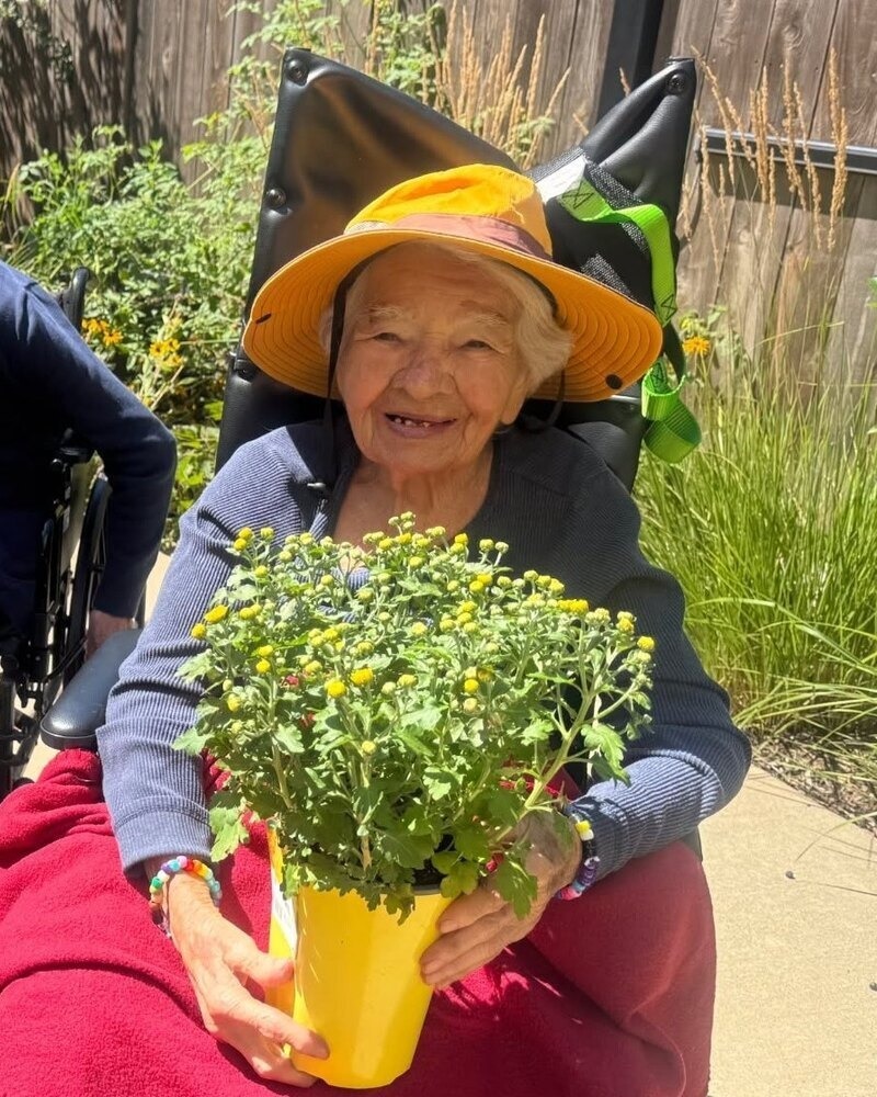 A smiling community member in a yellow sun hat holds a potted plant with yellow blooms while enjoying a sunny garden at Arbor Terrace Highland Park, where lush greenery reflects nature-friendly senior living Highland Park IL and warm senior care Highland Park Illinois.