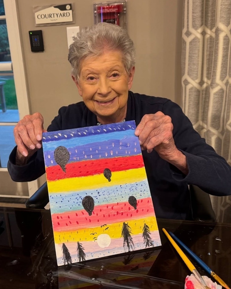 A smiling community member proudly holds up her colorful painting of hot air balloons drifting across a bright striped sky above silhouetted trees at Arbor Terrace Highland Park, one of the top senior living communities Lake County IL, celebrating an active senior lifestyle Highland Park IL.