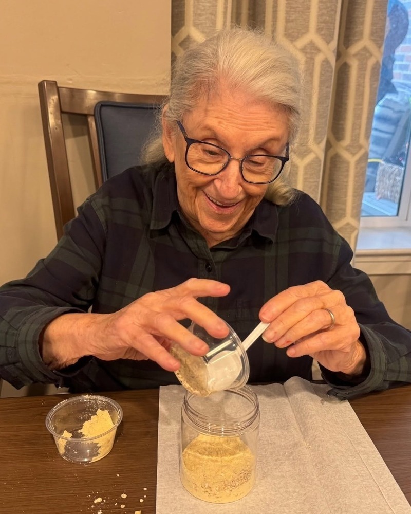 A smiling community member in a plaid shirt carefully pours a golden mixture from a small cup into a clear jar during a hands-on project at Arbor Terrace Highland Park, a cheerful moment that reflects an active senior lifestyle Highland Park IL and senior living Highland Park IL.