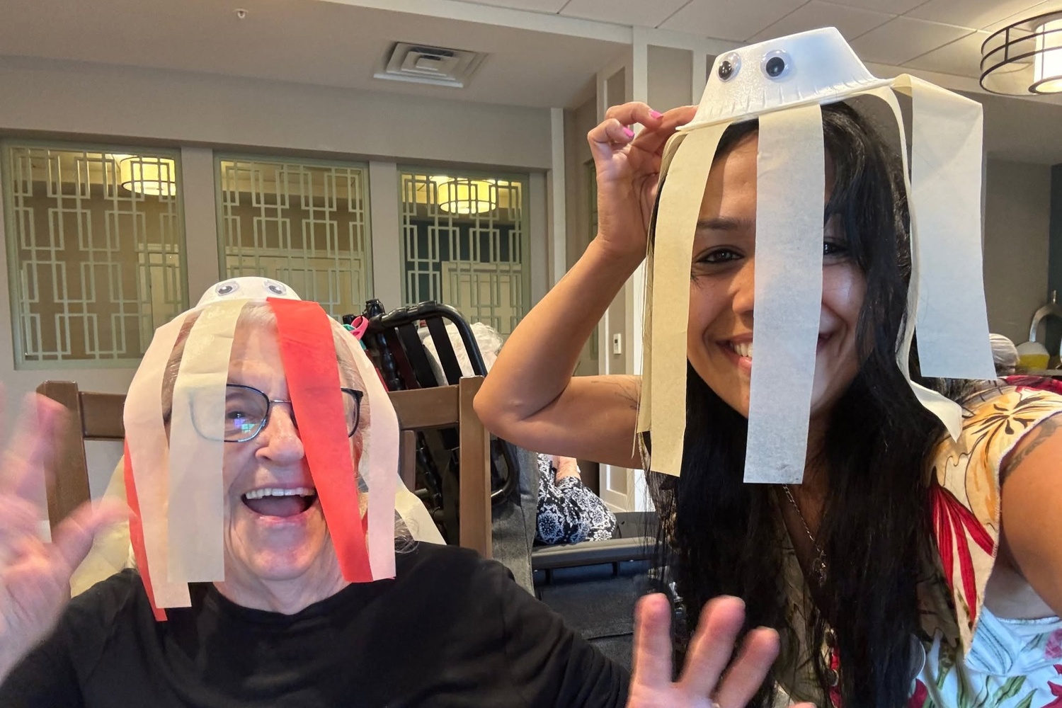 Two women laugh and wave while wearing playful paper plate hats with googly eyes and streamers at Arbor Terrace Highland Park, one of the top senior living communities Lake County IL, where creative crafts bring smiles to senior living Highland Park IL.