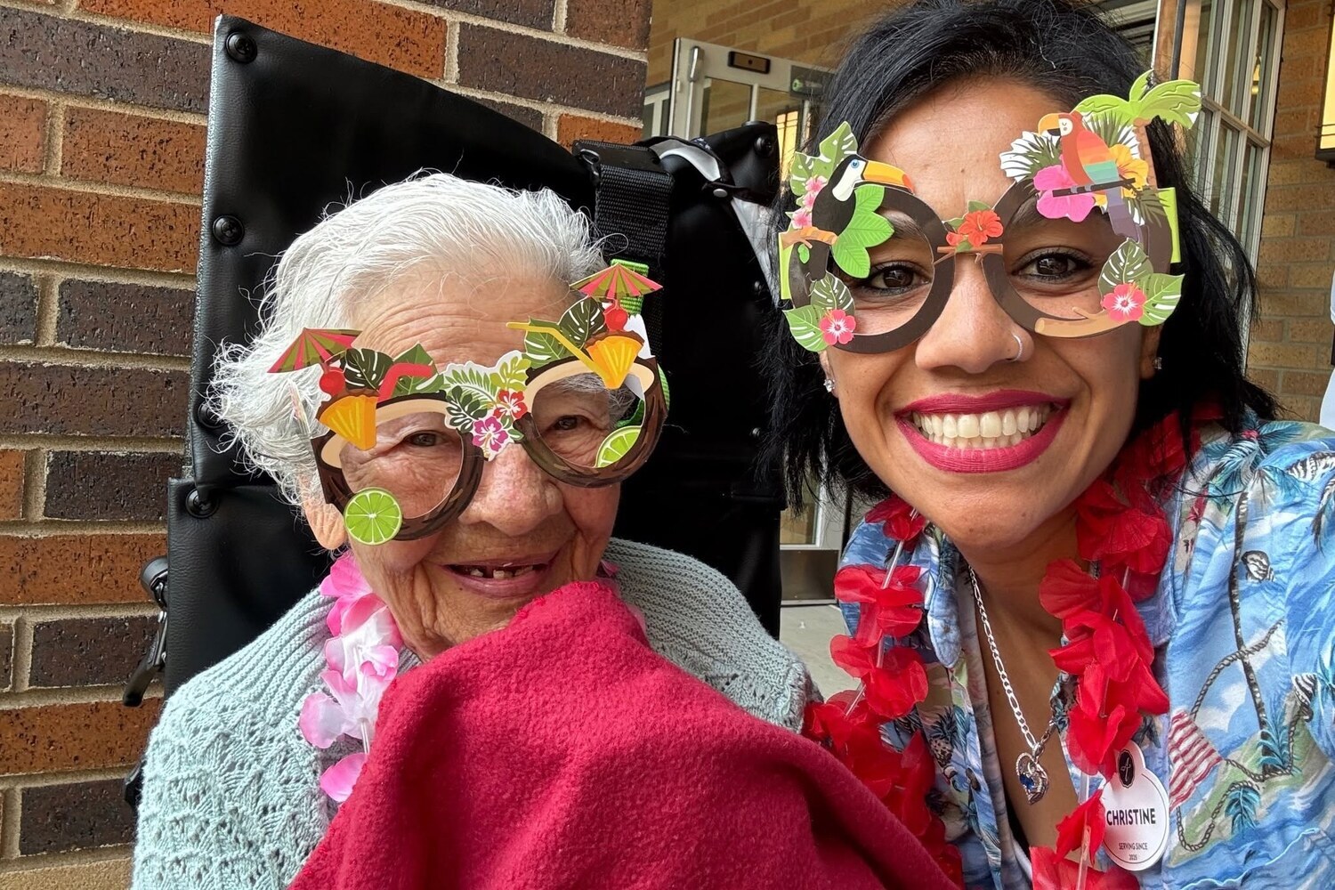 Two smiling women pose closely together in playful tropical-themed glasses and a cozy red wrap, a fun moment reflecting pet-friendly senior living Highland Park and resort-style senior living Lake County IL.
