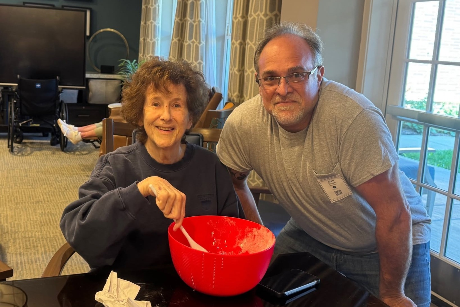 A smiling woman stirs a mixture in a bright red bowl as a man in a gray T-shirt leans in beside her, sharing a hands-on moment at Arbor Terrace Highland Park that reflects an active senior lifestyle Highland Park IL and welcoming senior care Highland Park Illinois.