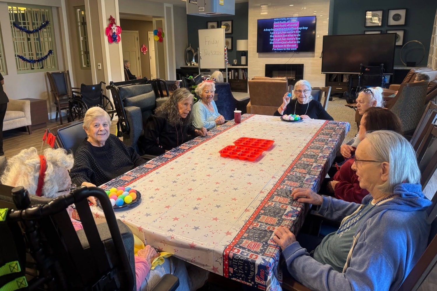 Community members gather around a long table draped in a festive tablecloth with red pieces stacked in the center, a screen glowing on the wall nearby, at Arbor Terrace Highland Park, a cozy group gathering that reflects senior living Highland Park IL and senior care Highland Park Illinois.