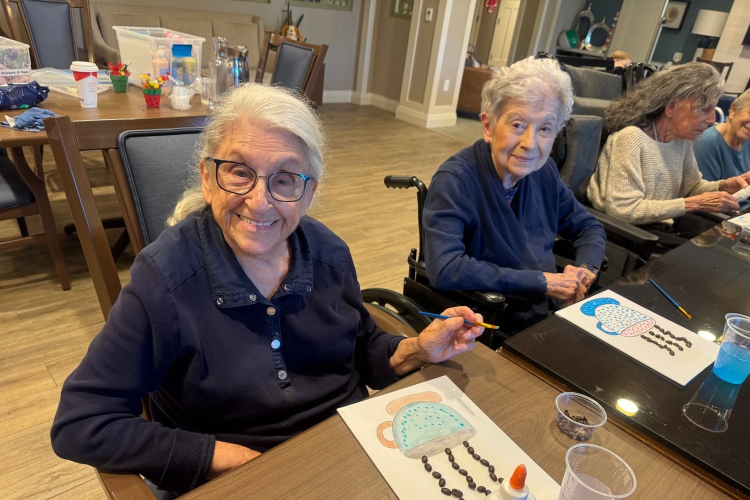 A smiling community member in glasses holds a paintbrush over a whimsical painting as others create their own artwork around a long table at Arbor Terrace Highland Park, one of the top senior living communities Lake County IL, where creativity is part of senior living Highland Park IL.