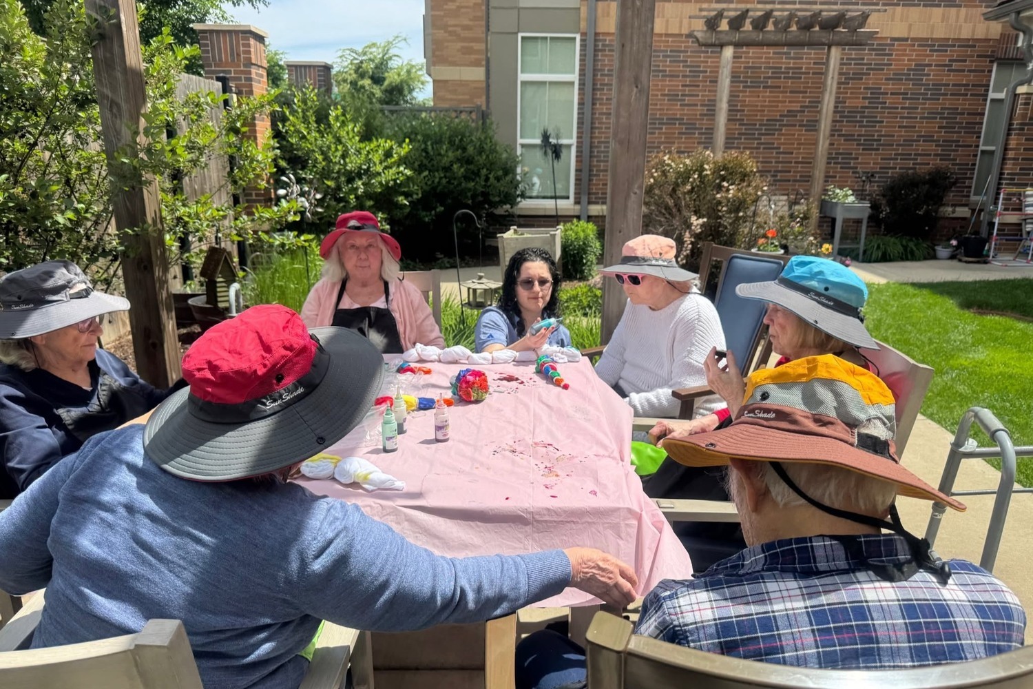 Community members in sun hats gather around a patio table with craft supplies, surrounded by leafy trees and garden paths at Arbor Terrace Highland Park, a creative outdoor moment that reflects senior living with walking paths Highland Park IL and senior care Highland Park Illinois.