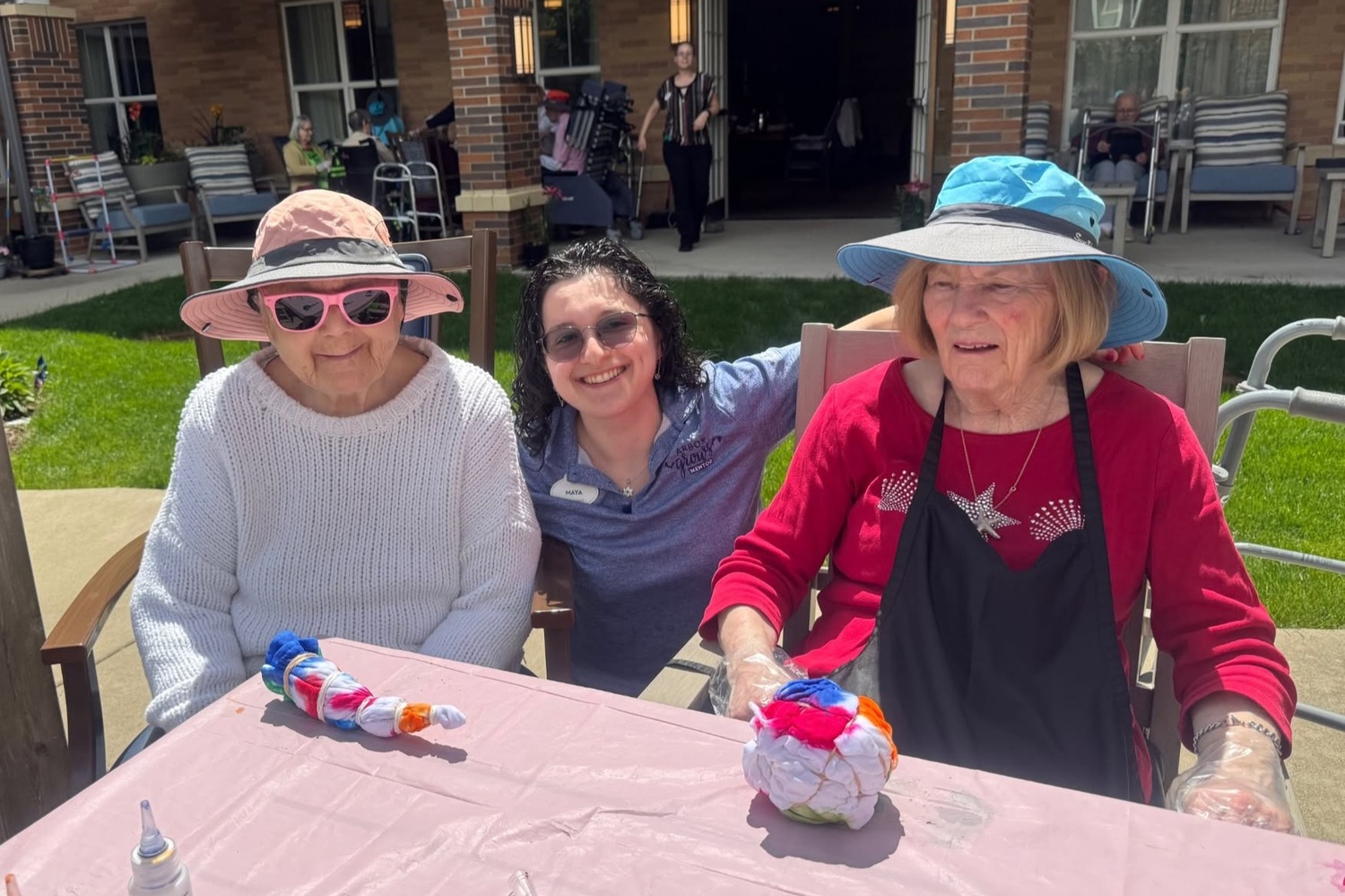 Three women in sun hats and sunglasses smile around a patio table with colorful tie-dye projects on a sunny day at Arbor Terrace Highland Park, where outdoor fun and green lawns reflect nature-friendly senior living Highland Park IL and an active senior lifestyle Highland Park IL.