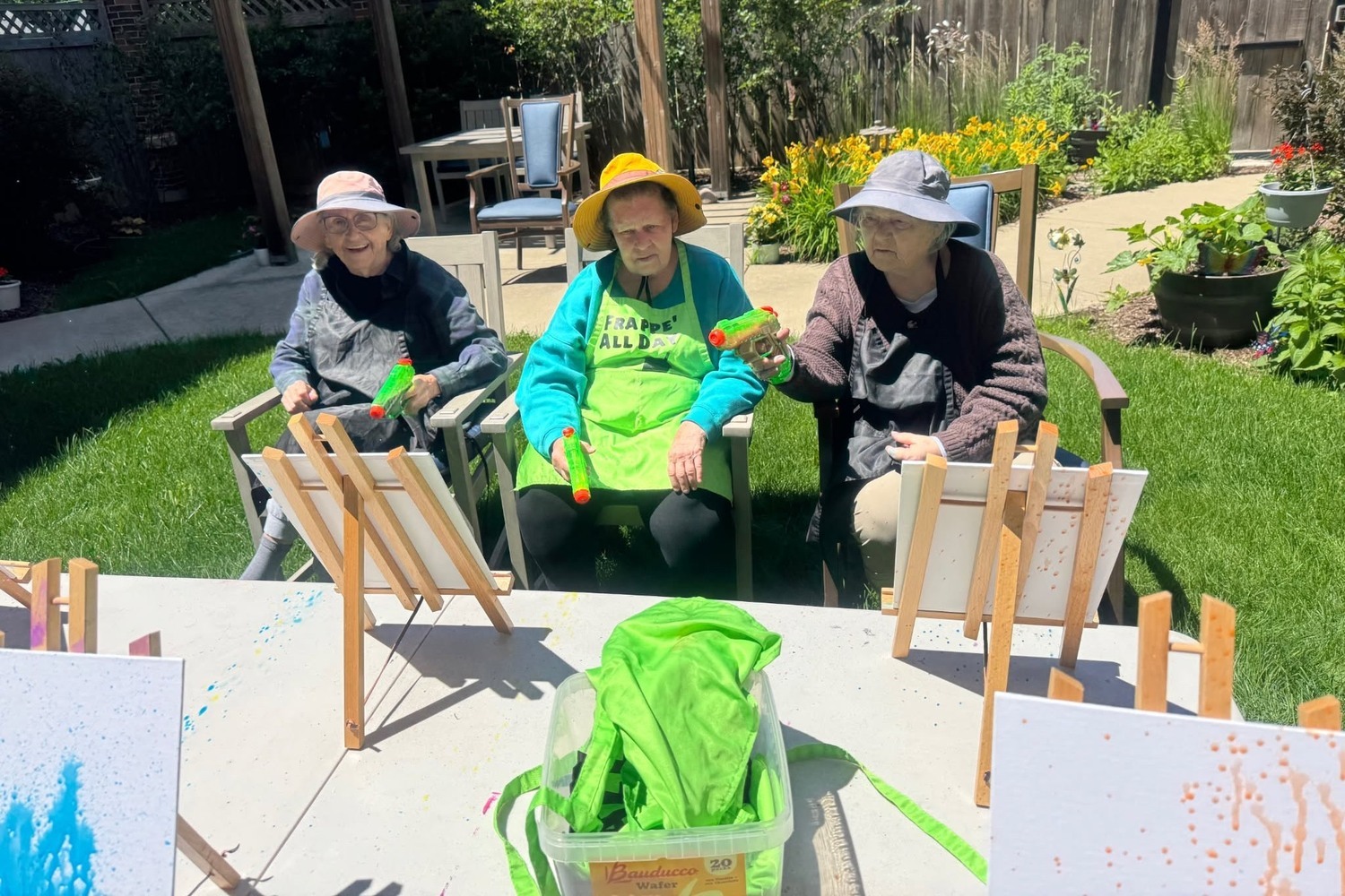 Three community members in sun hats hold colorful squirt guns beside easels on a sunny patio, with yellow flowers blooming in the garden behind them at Arbor Terrace Highland Park, a playful moment of nature-friendly senior living Highland Park IL and an active senior lifestyle Highland Park IL.