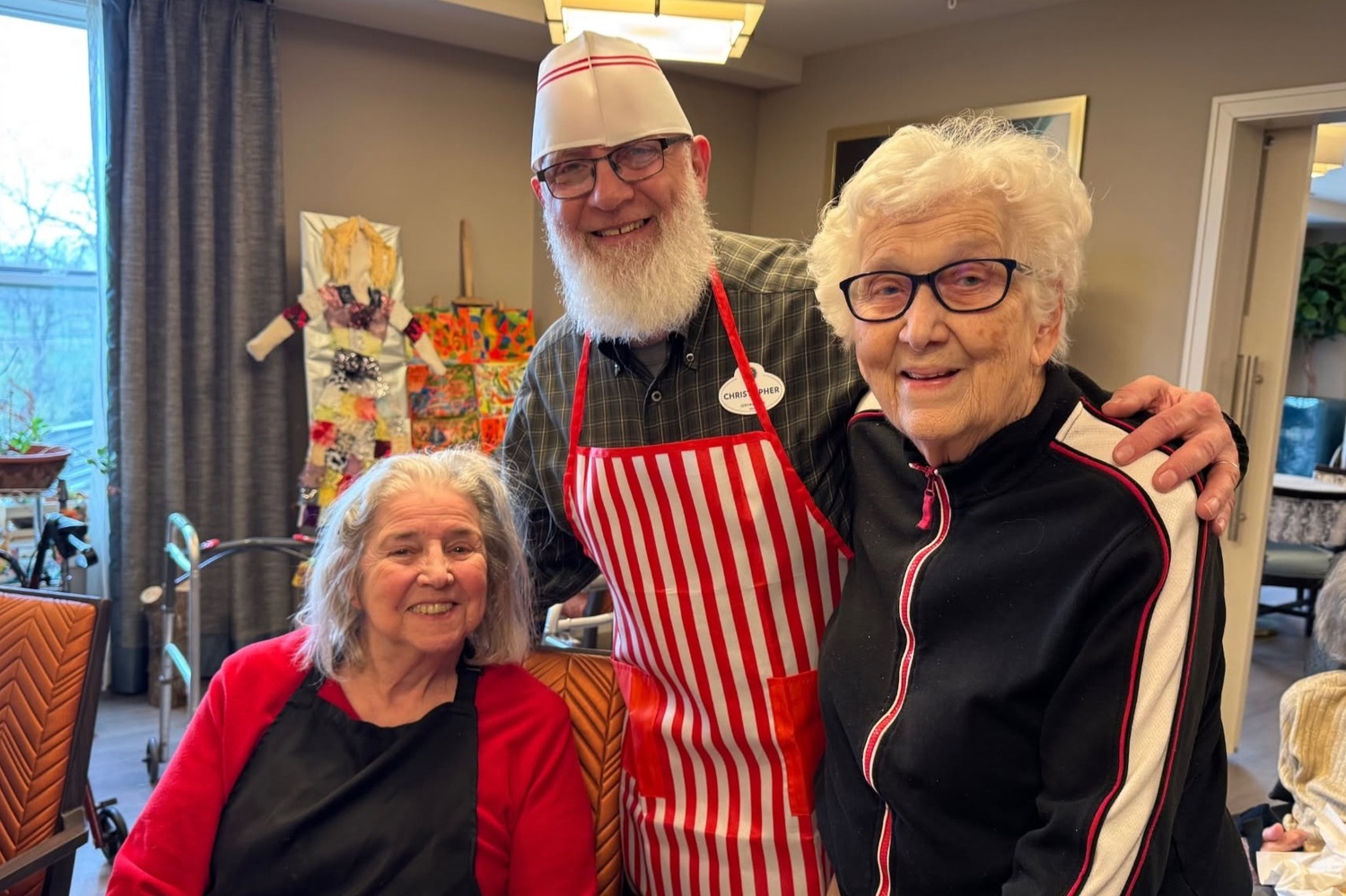 A bearded man in a white chef hat and red striped apron smiles with two women in front of colorful artwork at Arbor Terrace Highland Park, sharing the friendly spirit of senior living Highland Park IL and senior care Highland Park Illinois.