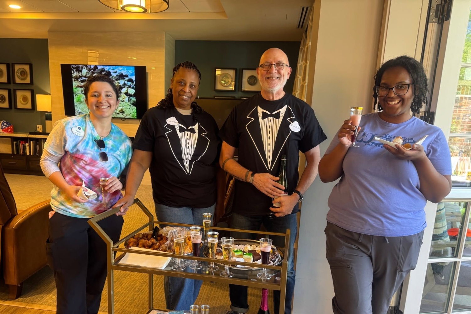 Four smiling people, two in playful tuxedo-print T-shirts, gather beside a gold bar cart filled with glasses and treats at Arbor Terrace Highland Park, one of the top senior living communities Lake County IL, where festive gatherings bring warmth to senior care Highland Park Illinois.
