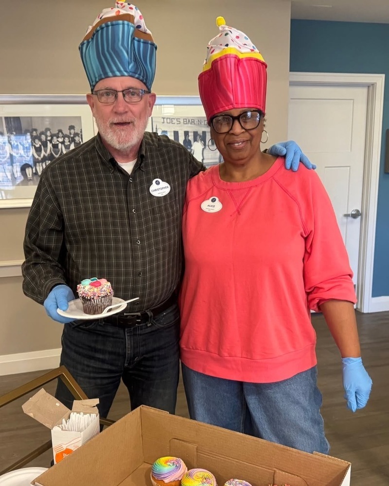 A man and a woman in playful cupcake hats smile side by side as he holds a chocolate cupcake on a plate, with a box of rainbow-frosted cupcakes ready to share at Arbor Terrace Highland Park, a sweet celebration of the active senior lifestyle Highland Park IL and senior living Highland Park IL.