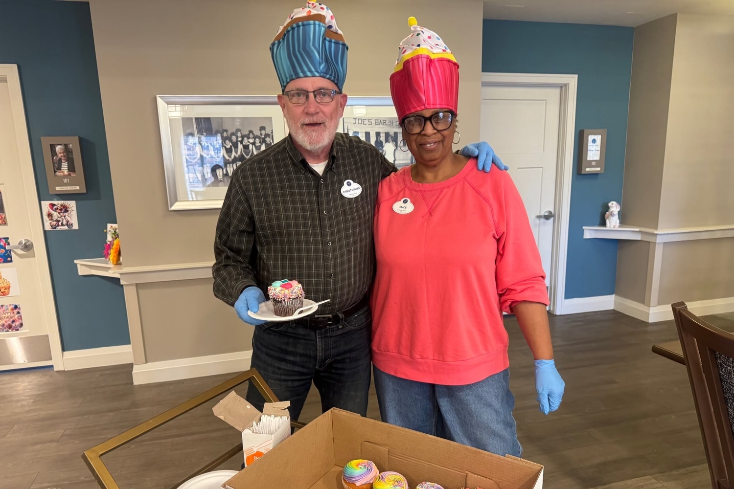 A man and a woman in playful cupcake hats smile together as he holds a frosted cupcake on a plate during a sweet celebration at Arbor Terrace Highland Park, where festive gatherings are part of senior living Highland Park IL and senior care Highland Park Illinois.