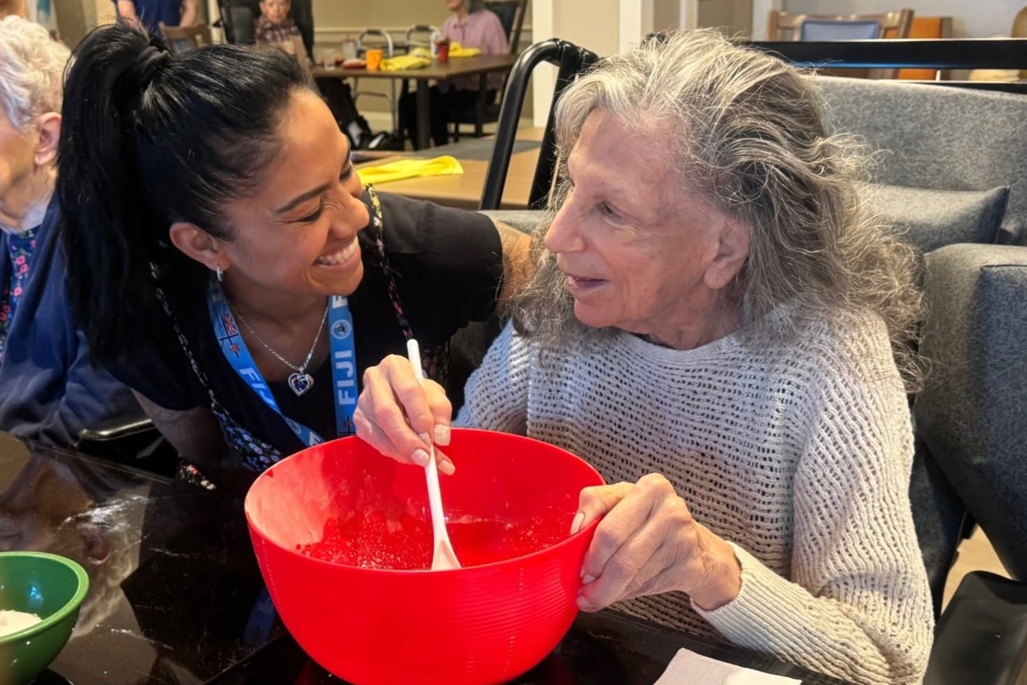 A smiling woman leans in close as a community member stirs a mixture in a bright red bowl, the two sharing a laugh at the table at Arbor Terrace Highland Park, a joyful hands-on moment that reflects an active senior lifestyle Highland Park IL and senior care Highland Park Illinois.