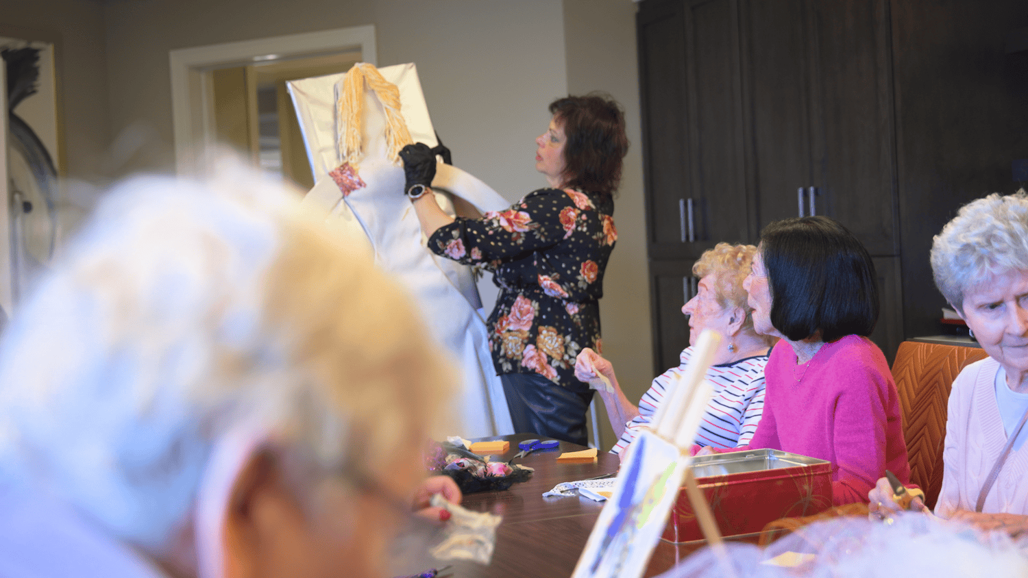 Residents gather around a table, smiling as a staff member dresses a cardboard cutout figure during a fun activity at our senior living community in Highland Park, IL.