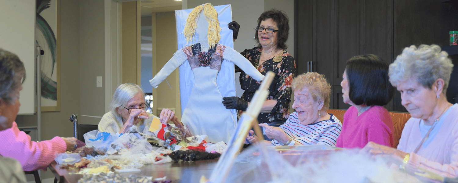 Senior women gather around a table covered in fabric and craft supplies as a woman in a floral top stands beside a tall handmade figure with golden yarn hair, a creative group project that reflects the active senior lifestyle Highland Park IL and senior living Highland Park IL.