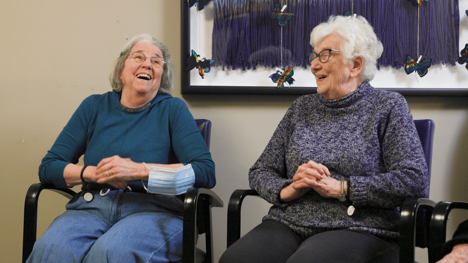 Two residents share a laugh together in front of hanging art in a colorful common area at our senior living community in Highland Park, IL.