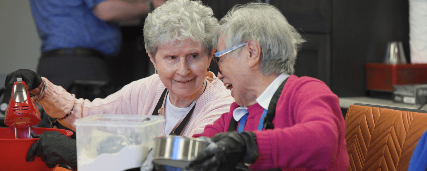 Two community members in aprons work side by side, mixing ingredients in red and black bowls during a shared hands-on activity at Arbor Terrace Highland Park, one of the top senior living communities Lake County IL, offering engaging senior care Highland Park Illinois.