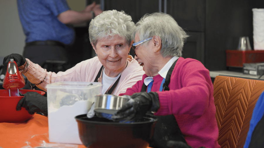 Two residents in pink and magenta cardigans laugh while mixing batter with black gloves and a red hand mixer during a baking activity at Arbor Terrace Highland Park. This reflects the active senior lifestyle and senior living in Highland Park, IL.