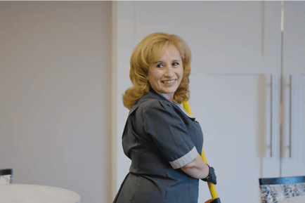 A smiling staff member in a gray uniform and dark gloves holds a yellow-handled cleaning tool and glances over her shoulder in a bright common area with white cabinetry, a welcoming glimpse of personalized senior care Highland Park IL and senior living Highland Park IL.