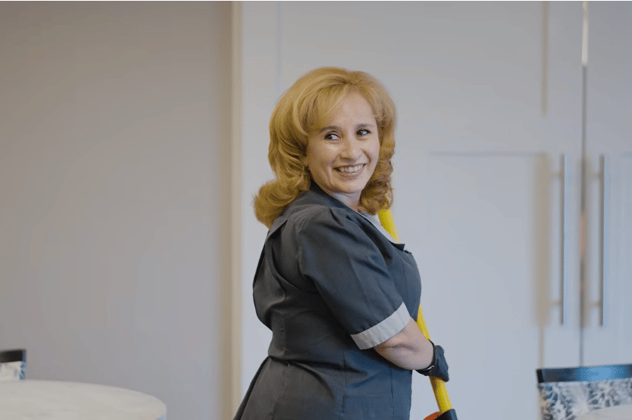 A smiling staff member in a gray uniform and dark gloves holds a yellow-handled cleaning tool and glances over her shoulder in a bright common area with white cabinetry, a welcoming glimpse of personalized senior care Highland Park IL and senior living Highland Park IL.