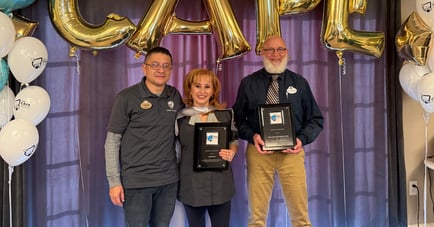 Three staff members smile while holding award plaques at an awards celebration at Arbor Terrace Highland Park, framed by gold balloon letters and white balloons. This reflects the dedication behind the senior living and senior care Highland Park, Illinois, families value.