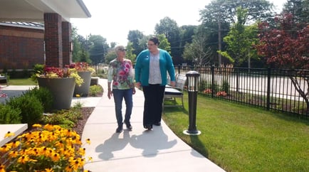 A resident and staff member stroll a flower-lined path at Arbor Terrace Highland Park, home to senior living with walking paths in Highland Park, IL. Nature-friendly senior living makes every outdoor moment special.