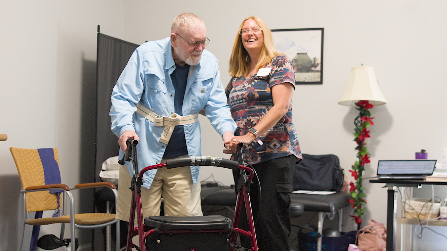 A smiling team member steadies a resident as he stands with a red rolling walker at Arbor Terrace Knoxville, in a room with a padded table and a laptop nearby, showcasing supportive senior care at this senior living community in Knoxville, TN.
