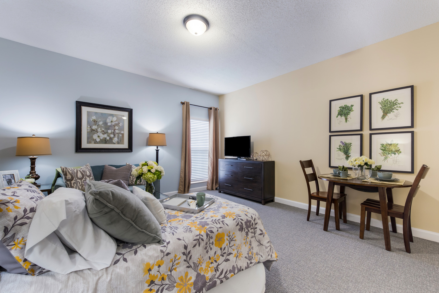 A model studio apartment at Arbor Terrace Knoxville with a breakfast tray on the bed, a flat screen TV on the dresser, and a bistro table set for two beneath framed plant prints, showcasing the comfortable apartments at this assisted living and memory care community in Knoxville, TN.