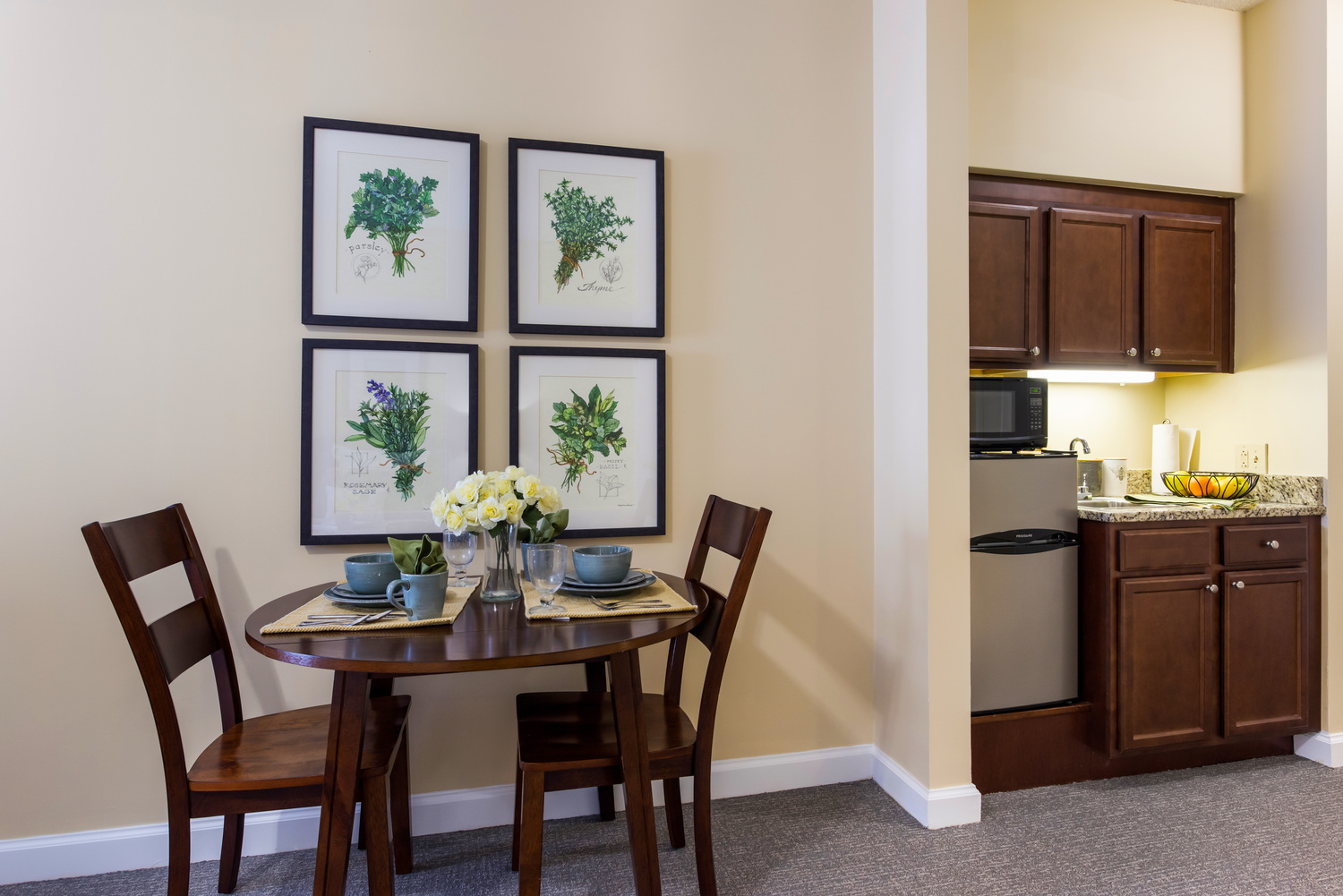 A dining nook and kitchenette in a model apartment at Arbor Terrace Knoxville, with a set bistro table under framed herb prints and dark cabinets with a microwave and small refrigerator, showcasing the apartments at this assisted living and memory care community in Knoxville, TN.