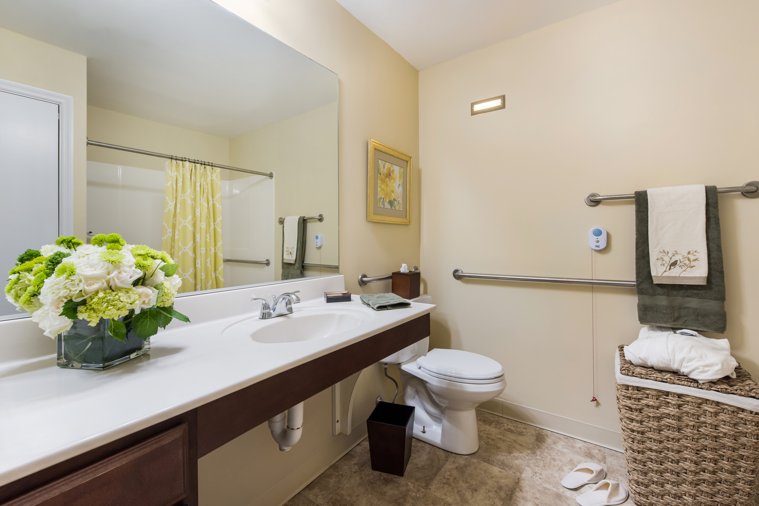 A model apartment bathroom at Arbor Terrace Knoxville with a wide vanity and mirror, grab bars, a call button, fresh flowers, and a robe and slippers set out, showcasing the thoughtful details at this assisted living and memory care community in Knoxville, TN.
