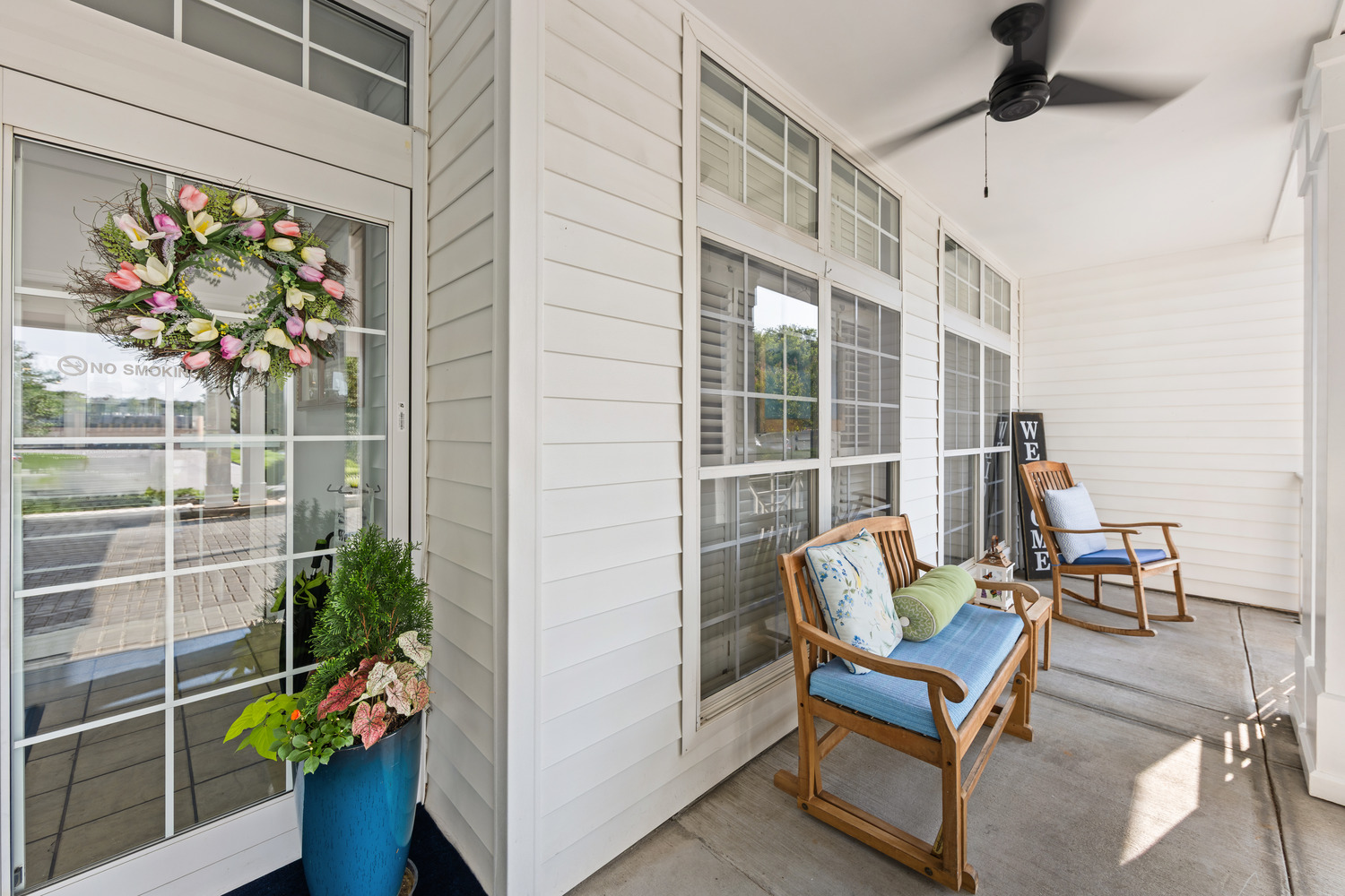 A front porch at Arbor Terrace Knoxville with a teak bench and rocking chair with blue cushions, a tulip wreath on the glass door, and a teal planter with greenery, showcasing the comfortable outdoor seating at this assisted living and memory care community in Knoxville, TN.